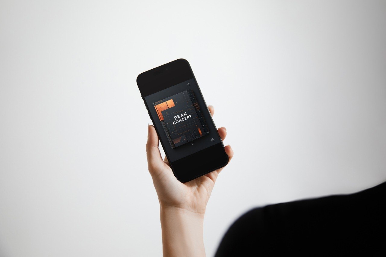 A hand holding a black smartphone that displays a digital cover or app interface with the text 'PEAK CONCEPT' in orange and white, set against a clean light gray background.