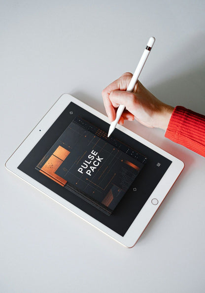 A white tablet on a light gray surface, with a hand holding a white stylus pointing at the screen. The screen displays a dark graphic template labeled 'PULSE PACK' with orange geometric shapes.